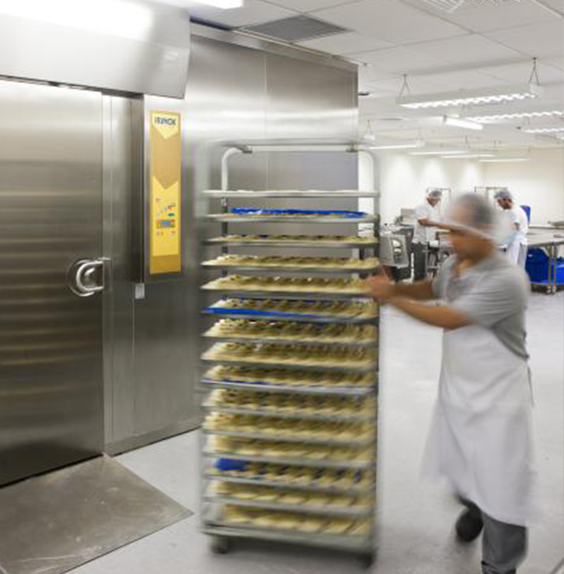 A baker in a controlled enviroment pushing a trolley of pastrys into a cold room